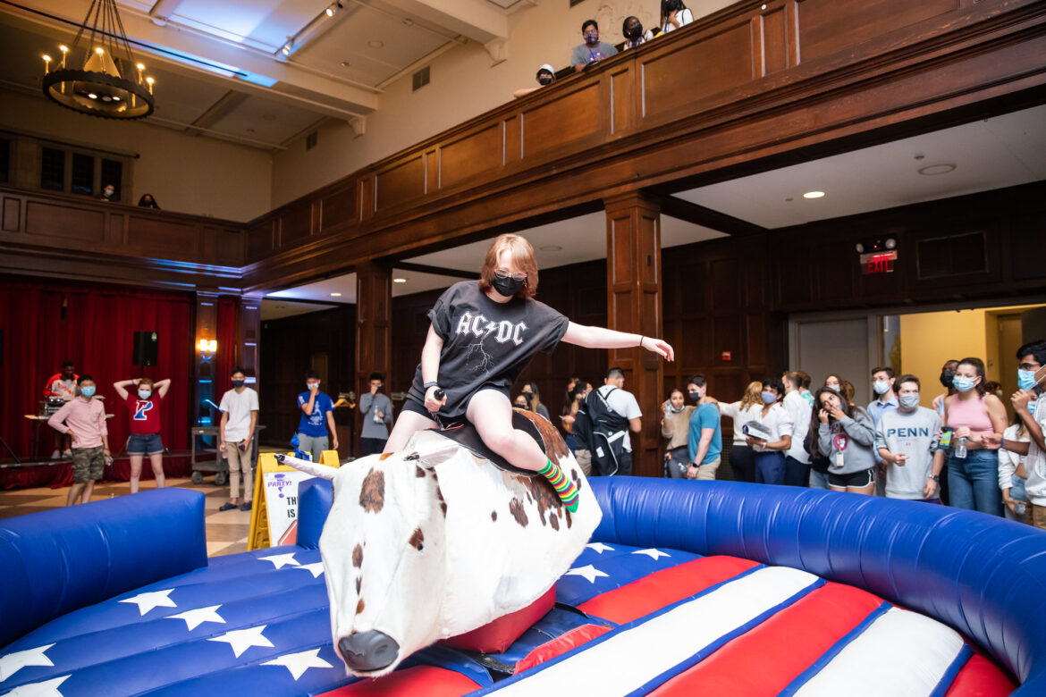 Student riding a mechanical bull in a ring with the pattern of the American Flag in the Hall of Flag room with students watching. 