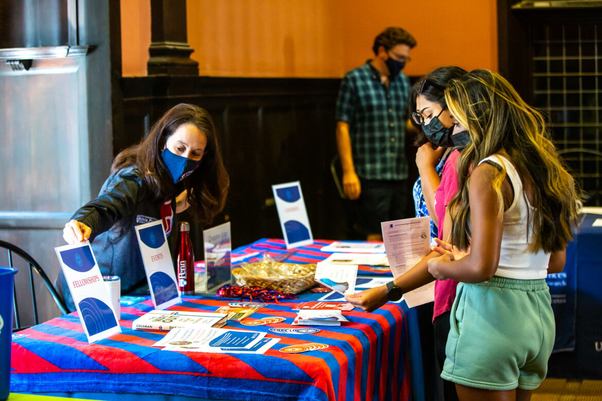 One student and one parent at a Penn Table . The table has red and blue stripe table cloth and three blue and white signs with a women behind the table.
